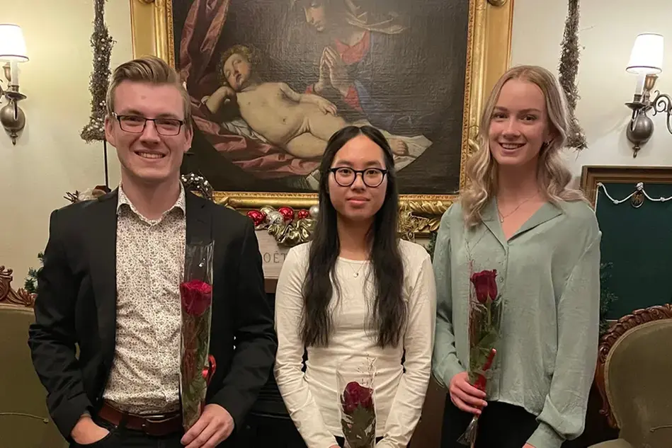Tre finklädda studenter håller i en varsin ros och ler mot kameran. I bakgrunden hänger en renässanstavla och vägglampor.