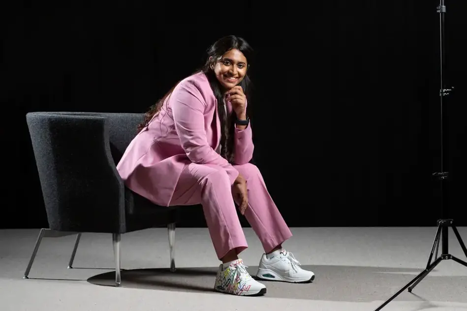En kvinna i en rosa kostym sitter på en stol och ser in i kameran. Foto. 