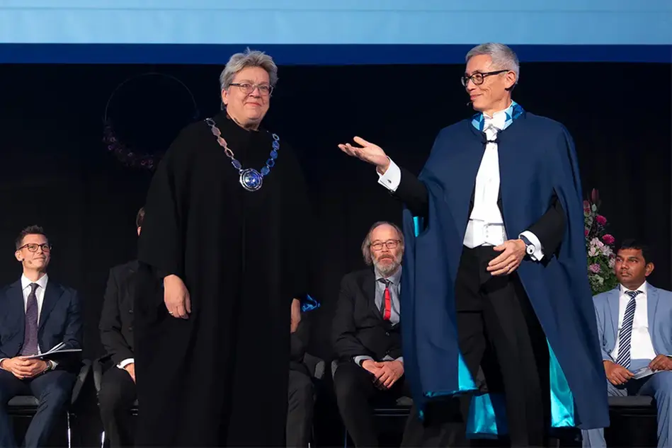 A woman with a black gown and a large piece of silver jewelry around her neck stands next to a man in a black gown. They are both smiling. Photo.