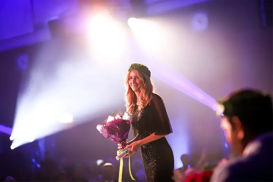 A woman with light long hair and a dark, glittering dress stands on a stage. In her hands she holds a bouquet of flowers and on her head she has a wreath. Photo.