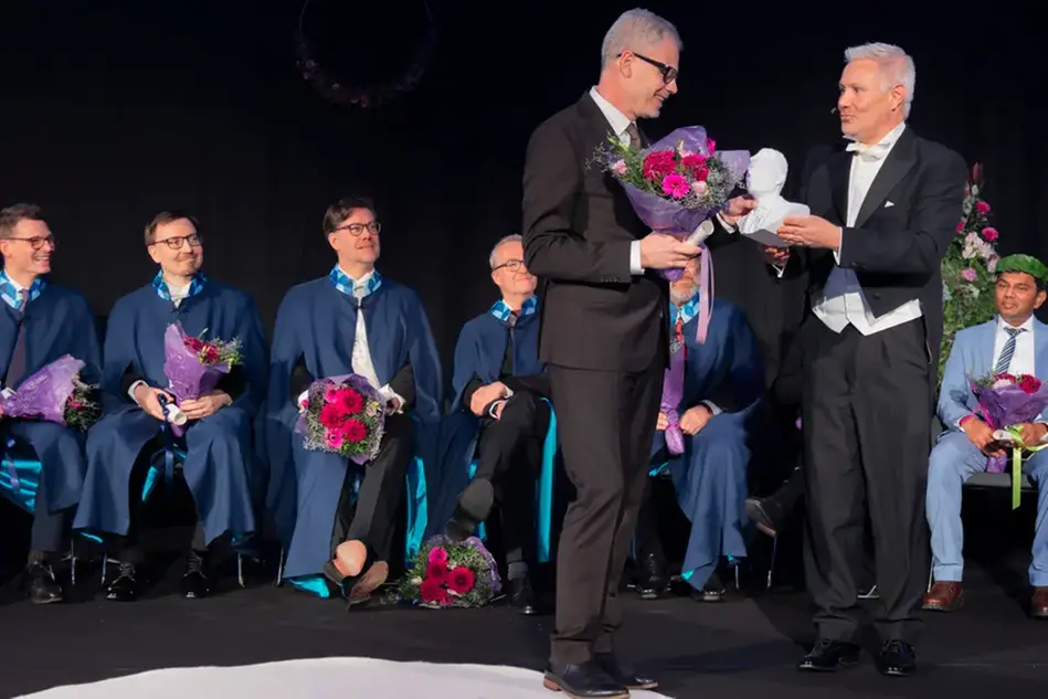 Two people dressed up on a stage. One is giving the other a prize and a bouquet of flowers. Several people are sitting in the background. Photo.