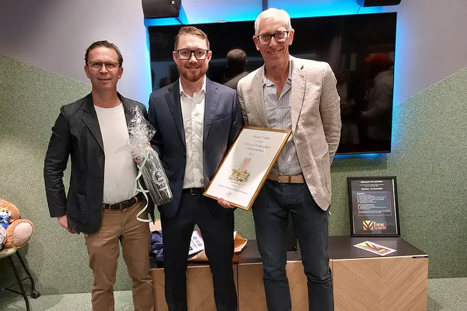 Tre män står framför en tv. Mannen i mitten håller upp present och diplom. Foto.