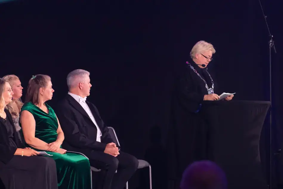 A short-haired woman in a black outfit is standing at a podium on a stage and gives a speech. A row of dressed up people are sitting on the stage.