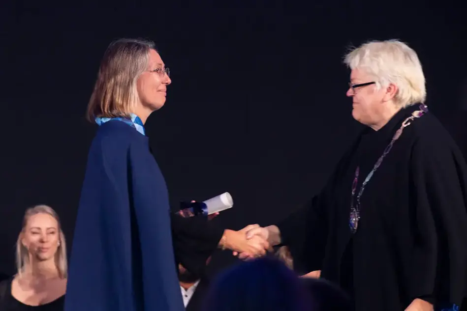 Two women stand on a stage and shake hands. One of the women has a diploma in her hand.