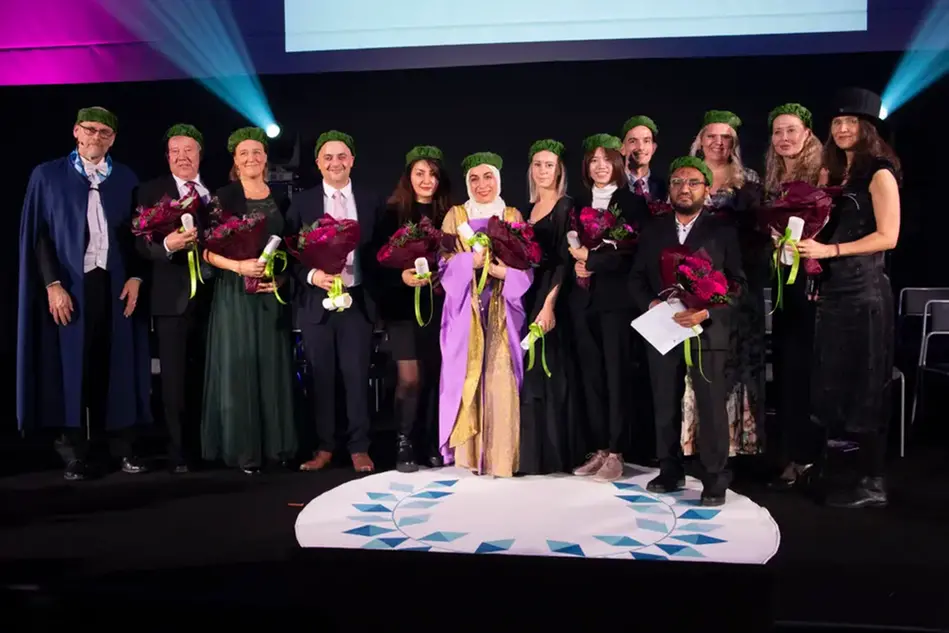 A stage with well-dressed people in a row. Several of them have wreaths on their heads and diplomas in their hands.