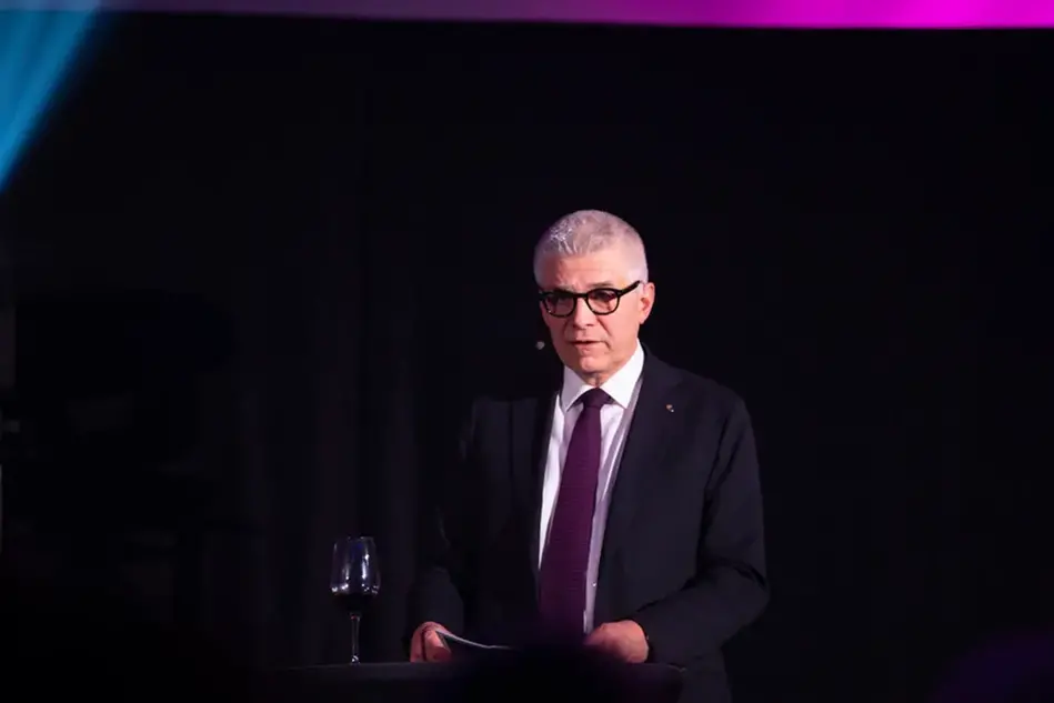 An elderly man, wearing glasses, in a black suit stands at a podium on a stage and gives a speech.