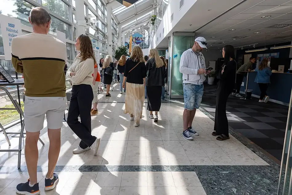 Många personer i en gång med glasväggar och glastak minglar. Flera personer står vid höga bord och berättar om sin verksamhet för besökare. Foto.