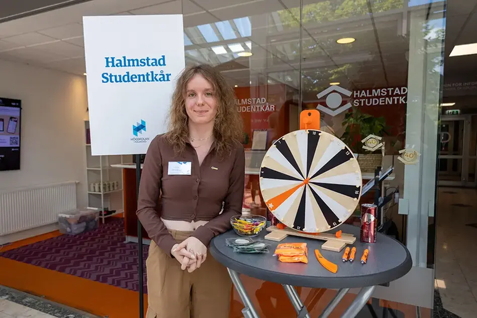 En ung person står vid ett bord med olika tävlingsprylar på. Personen ler och bakom syns texten "Halmstads Studentkår". Foto.