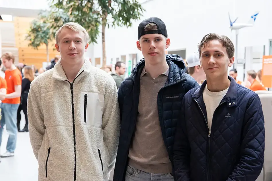 Tre unga män står uppdarade bredvid varandra i en ljus lokal. I bakgrunden syns ett vimmel av människor.