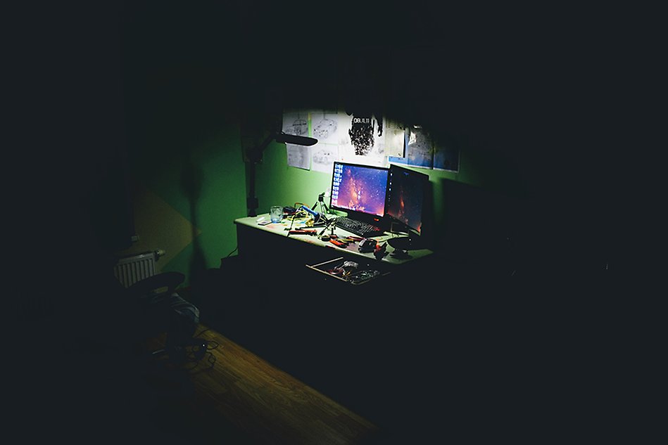 A dark room with a dimly lit desk with several computer monitors on it. Photo.