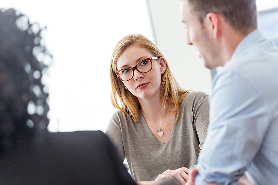 A young woman sitting down conversing with two people with their backs towards the camera