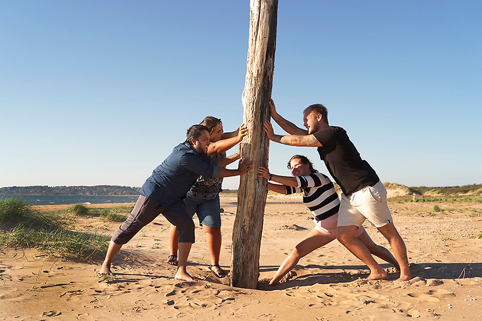 Four people holding a log upright on a beach. Photo.