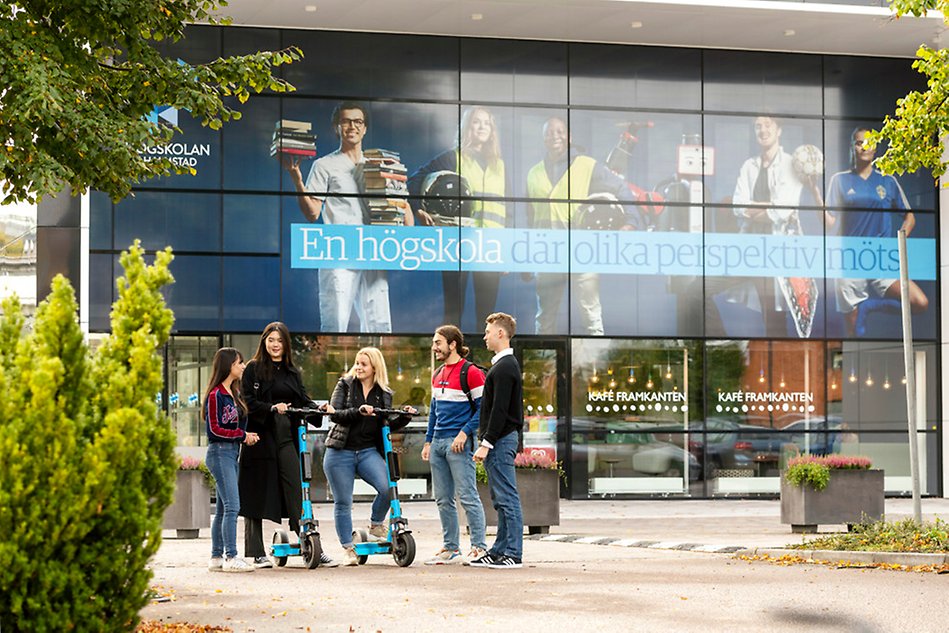 Tre tjejer och två killar står utanför huvudentrén till Högskolan i Halmstad. Två av tjejerna har varsin sparkcykel. Foto.