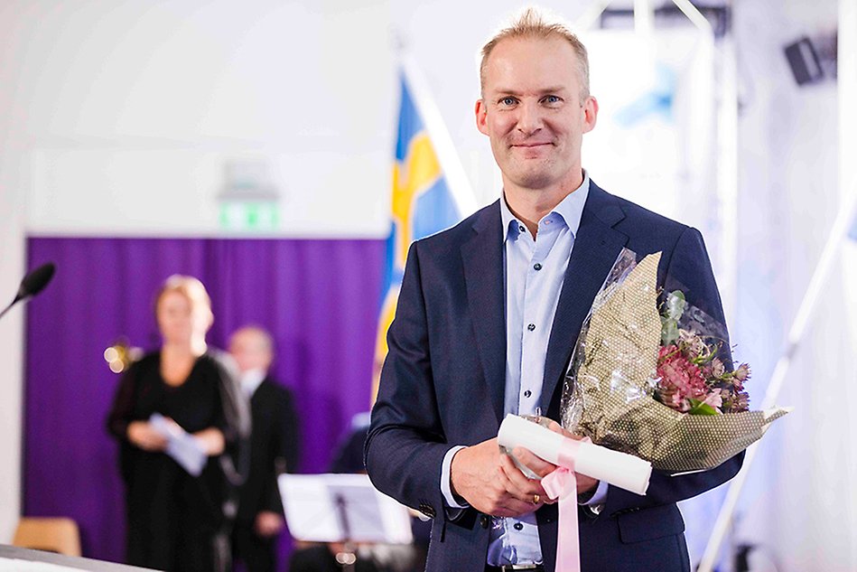 Man holding flowers, a diploma and a glass apple, looking into the camera and smiling. Photo.