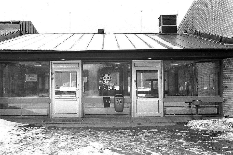 Black and white close-up of a building with two entrance doors. The previous University logo is displayed on a window in the center. Photo.