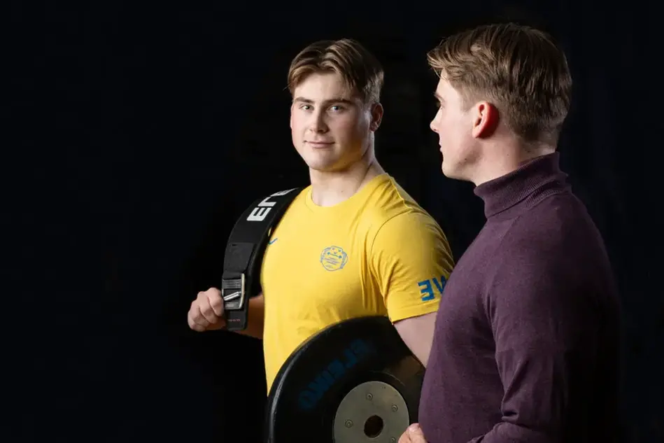 En man i vinröd polotröja tittar på sin spegelbild. Hans spegelbild har på sig en gul träningst-shirt och håller i en viktplatta och ett lyftarbälte. Foto.