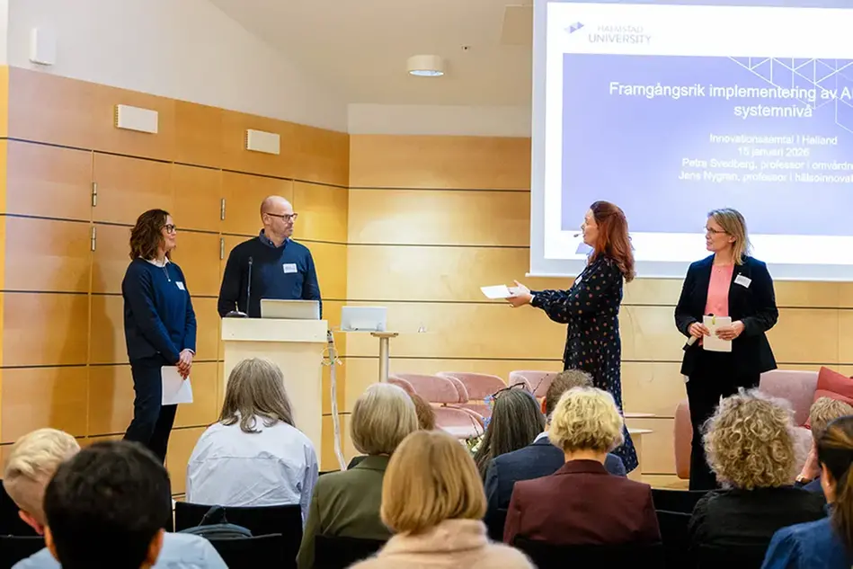 Fyra personer står vid ett podium framför en publik. I bakgrunden en presentation. 