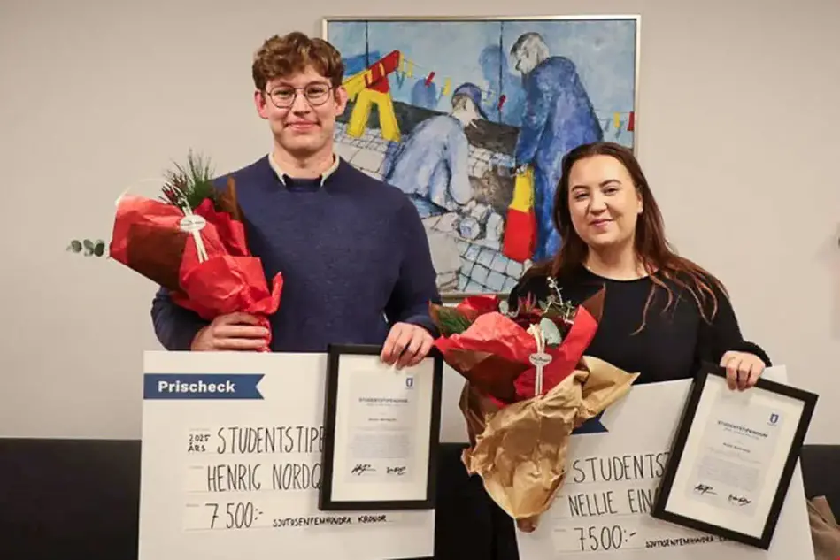 Två studenter ler mot kameran. De håller i varsitt diplom, bukett och värdecheck. Bakom hänger en färgglad tavla på en vit vägg.