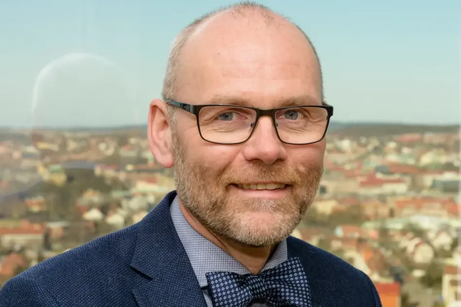 En man med glasögon och blå kostym står framför en stadsbild. Foto