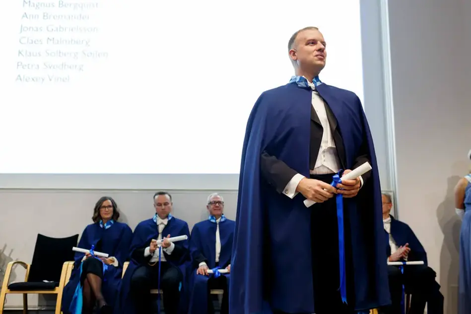 En man klädd i en blå cape och med ett diplom i handen står framför en projektion på skärmen. En grupp andra människor i caper sitter bakom mannen. Foto 