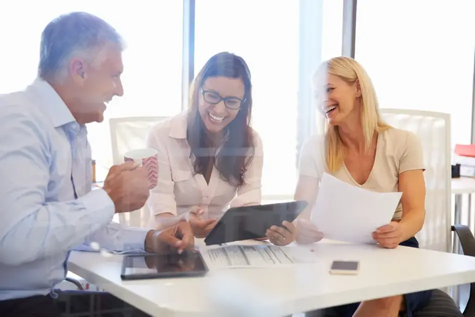 Three people sits around, integrates with each other, with a table with computers and papers on it.