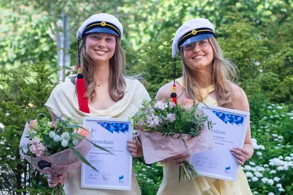 Två studenter i hatt och gul klänning ler mot kameran. De håller i diplom och blombukett. I bakgrunden finns grönska.