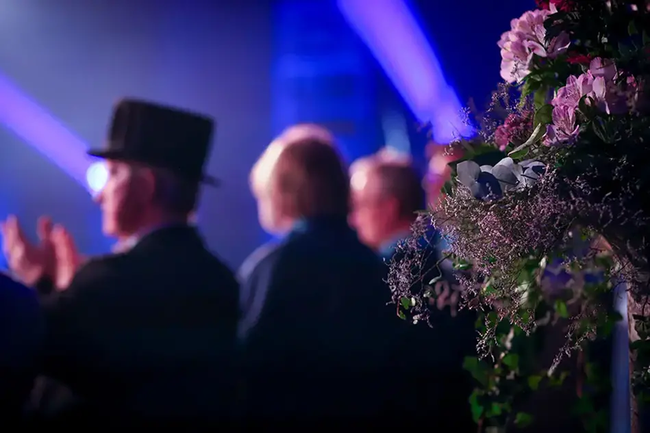 Oskarpa personer i mörkt ljus, med en stor bukett blommor i förgrunden. En person bär en svart hatt.