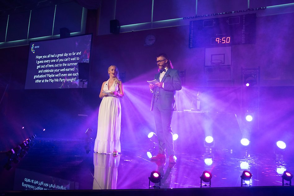 En kvinna i klänning och en man i kostym står på en scen med ljusa bakgrundslampor. Foto.