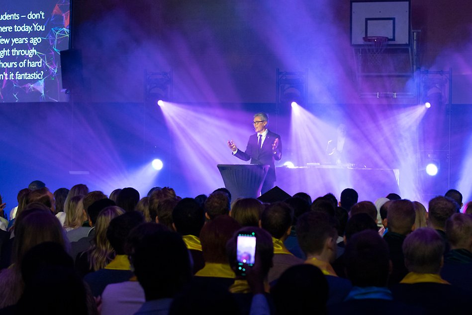 A man is standing on a stage by a podium giving a speech. In front of him is an audience. Photo.