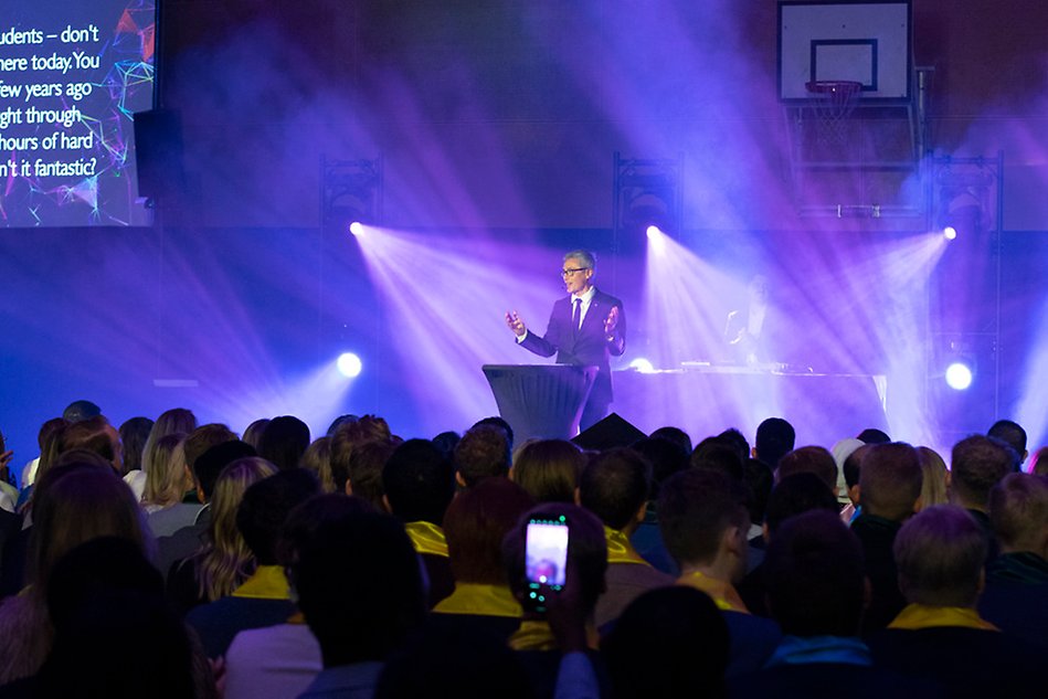 En man står på en scen vid ett podium och håller tal. Framför honom syns ett publikhav. Foto.