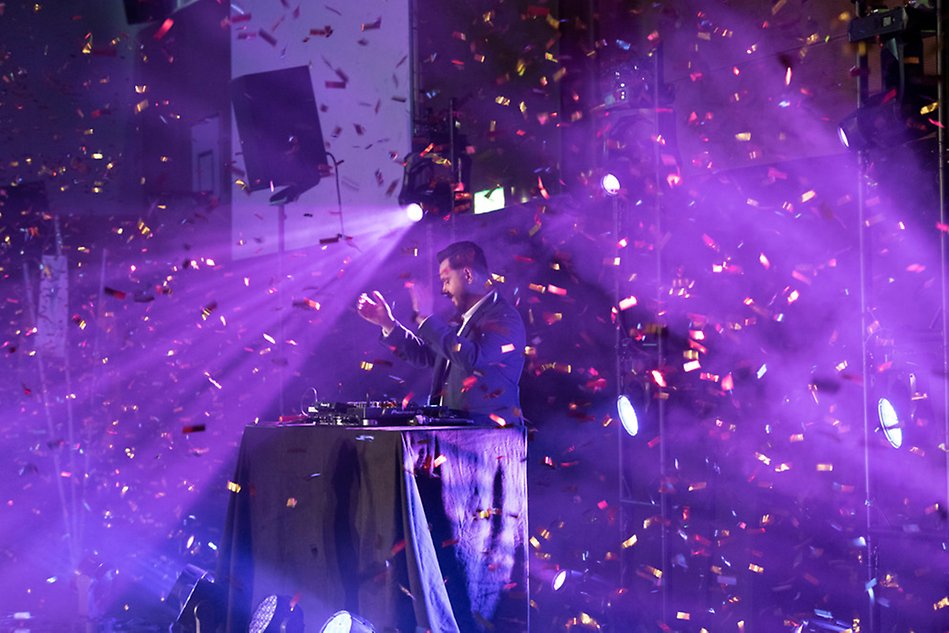 A discjockey is standing on a stage by a mixer table. Confetti is raining down. Photo.