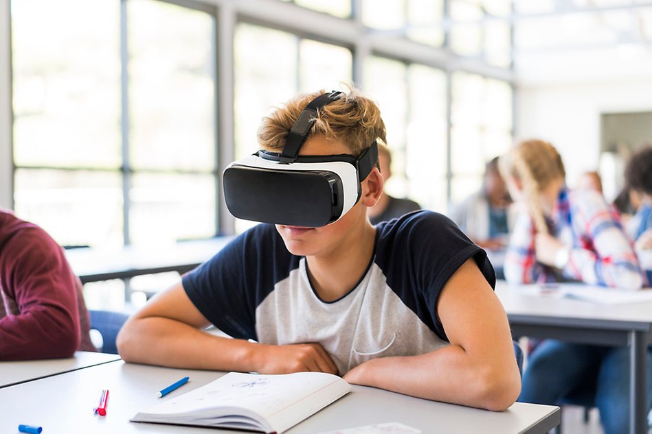 A boy wearing virtual reality glasses sits at a desk in a classroom. Photo.