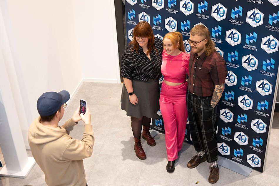 Three people are being photographed in front of a photo wall by a person with a cap. Photo.