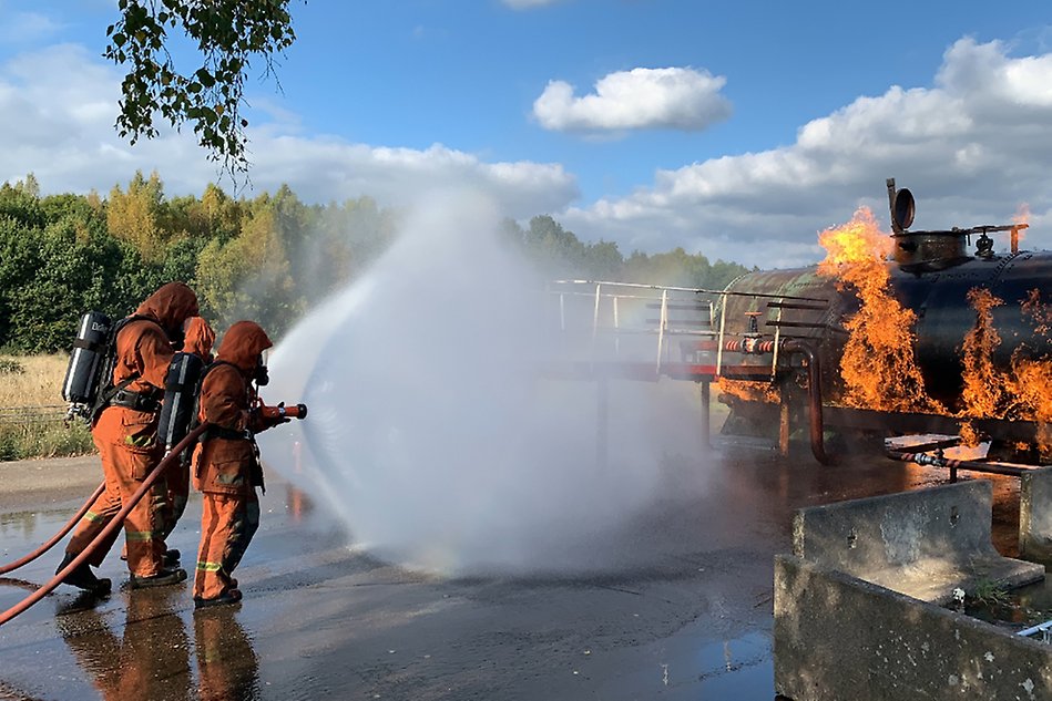 Two people wearing safety gear are putting out a vehicle fire.