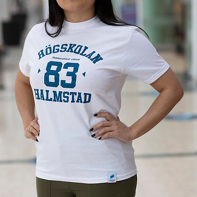 En vit t-shirt med texten ”Högskolan 83 Halmstad” i blå text sitter på en person vars överkropp syns i bild. Foto.