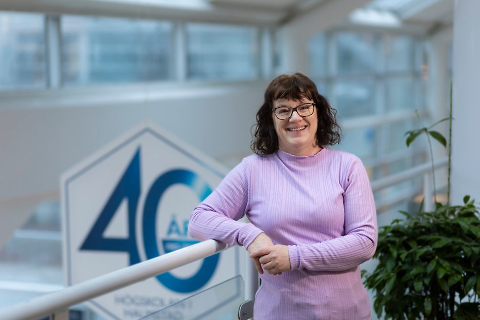 Woman with glasses and brown hair is standing against a railing. Kvinna med glasögon och brunt hår står lutad mot ett räcke. Photo.