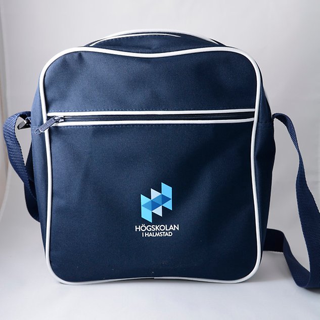 En mörkblå väska med axelband och Högskolan i Halmstads logotyp sedd framifrån. Foto.