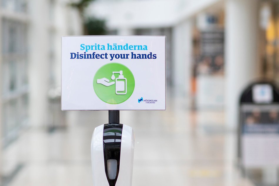 En skylt i förgrunden uppmanar till att sprita händerna mot en suddig bakgrund.