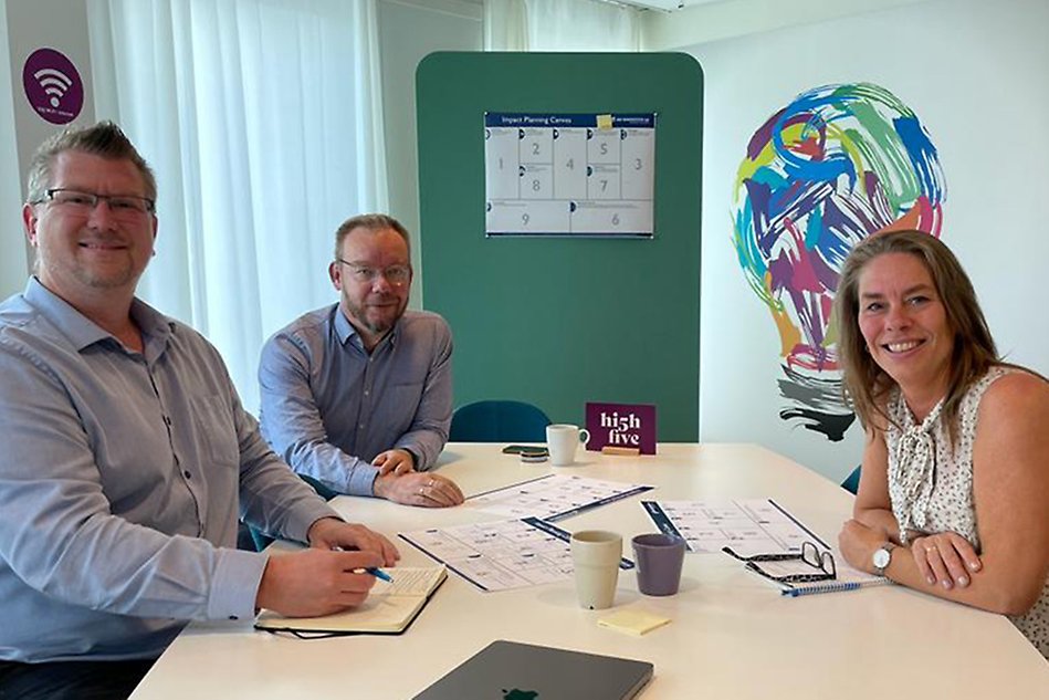 Three people sitting by a table with papers on it, looking into the camera. Photo.