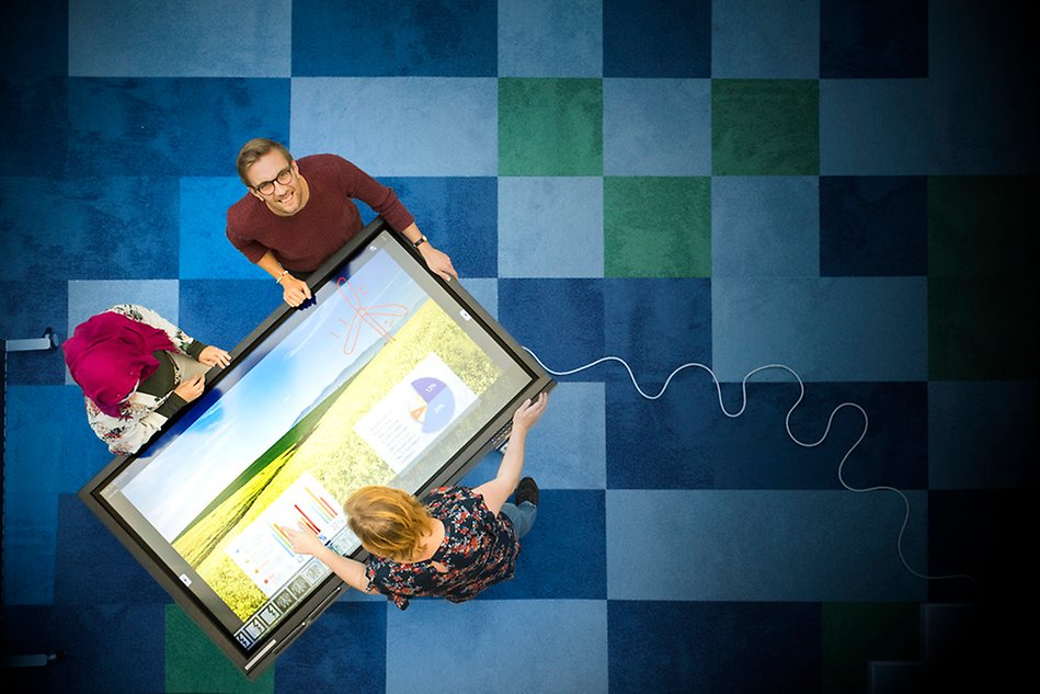Rutig matta, tre personer runt en stor digital skärm som tippats till liggande läge. En person tittar upp mot kameran. Foto.