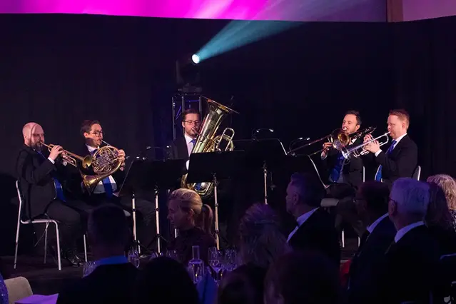 En orkester sitter på en scen och spelar. I förgrunden skymtar delar av publik.