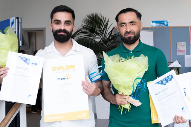 Två studenter i pikétröja håller upp diplom och blommor. Foto.