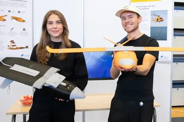 Två studenter ler mot kameran. De håller i en varsin modellflygplan. Bakom dem är en monter.