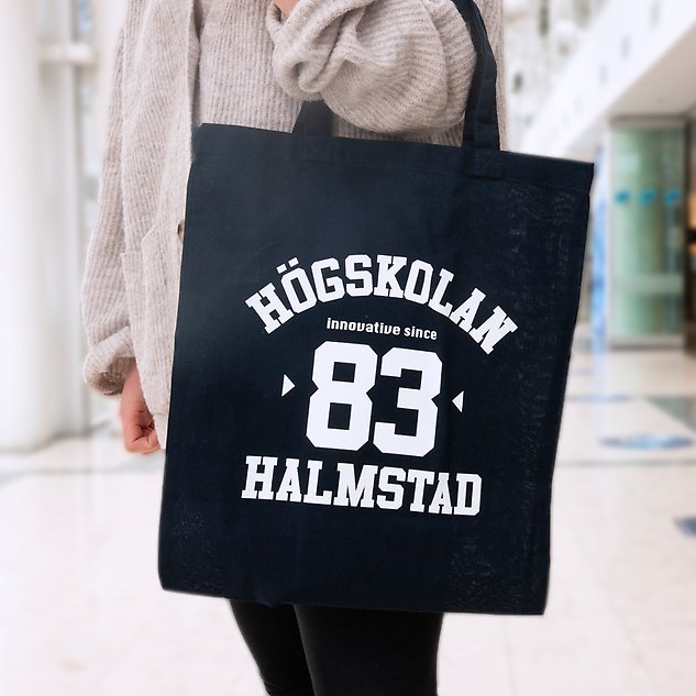 A dark blue tote bag with the text ”Högskolan 83 Halmstad” hangs on the armof a person seen from the side. Photo.