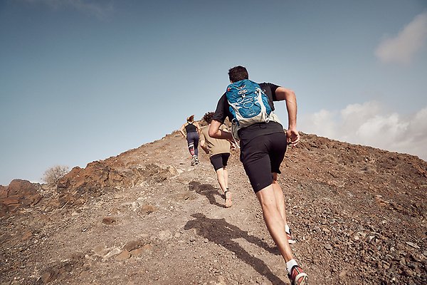 Tre löpare med ryggen mot kameran springer på rad uppför ett kargt, stenigt berg. Foto
