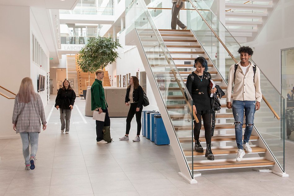 Två personer går ned för en trappa i en ljus och rymlig trapphall. Nedanför trappan står två personer och samtalar med varandra. Foto.