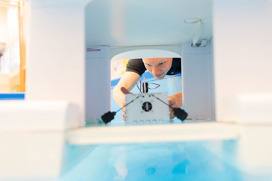 A young man seen through a tunnel is lowering a small vessel into a pool. Photo.