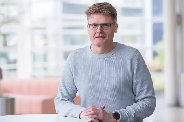 Person i grå tröja med glasögonen och kort hår som ler mot kameran
