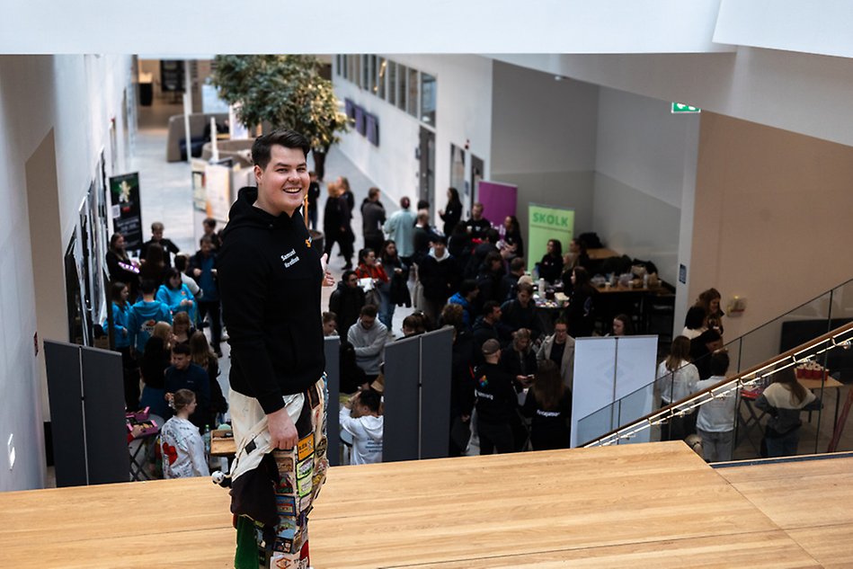 Person in a black jumper wearing student's overalls. He is standing in front of people at a fair. Photo.