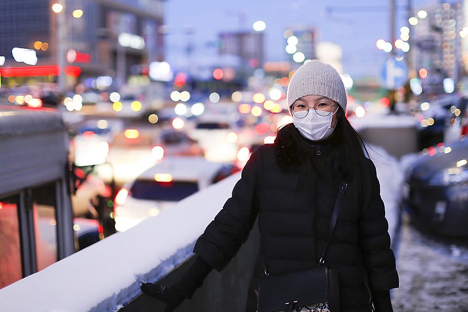 A woman in glasses and a face mask standing outside wearing a thick jacket and a beanie, city lights and traffic in the background. Photo.